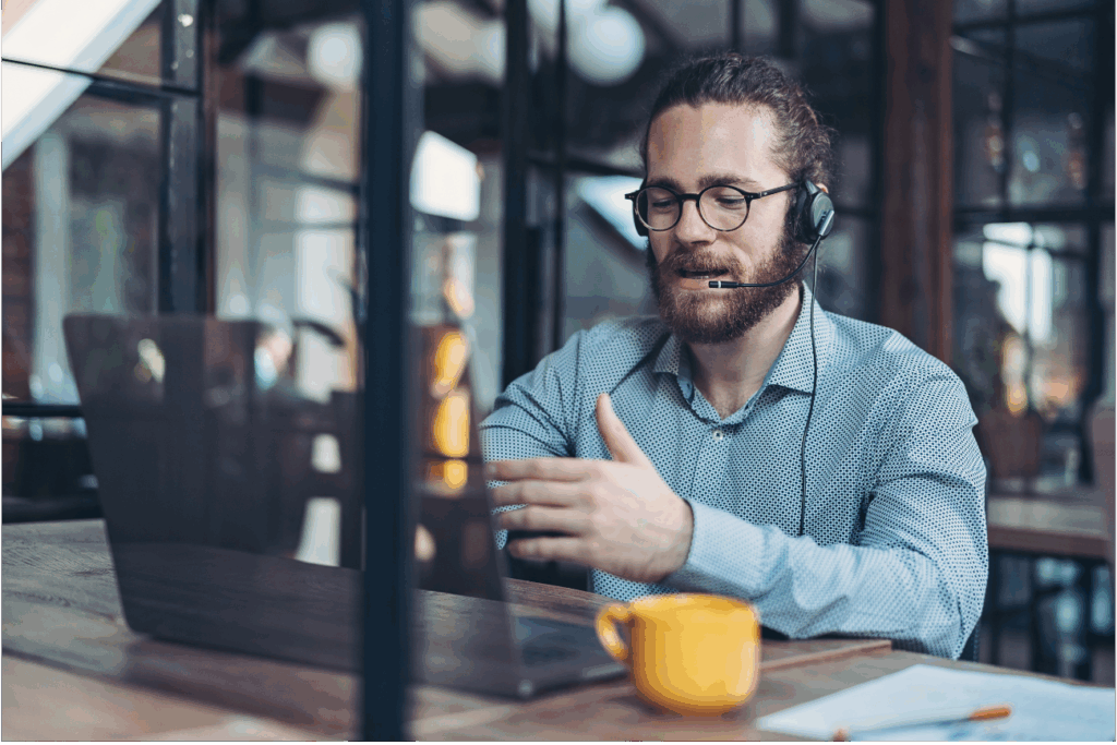 A professional who is working on a laptop, wearing a headset and appears to be talking. Orange mug on the table