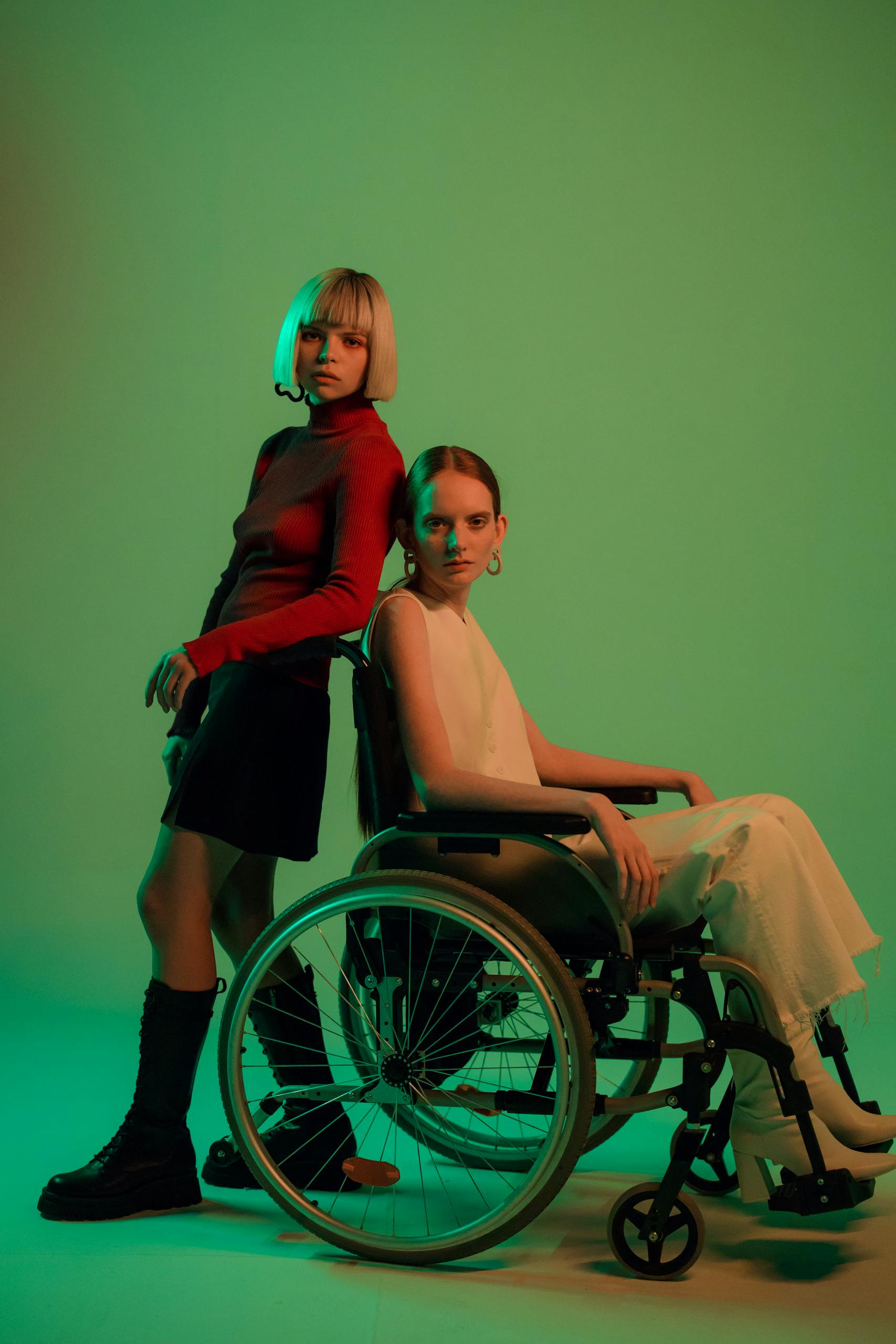 Two women in a stylish studio setting, one in a wheelchair, against a light green background.